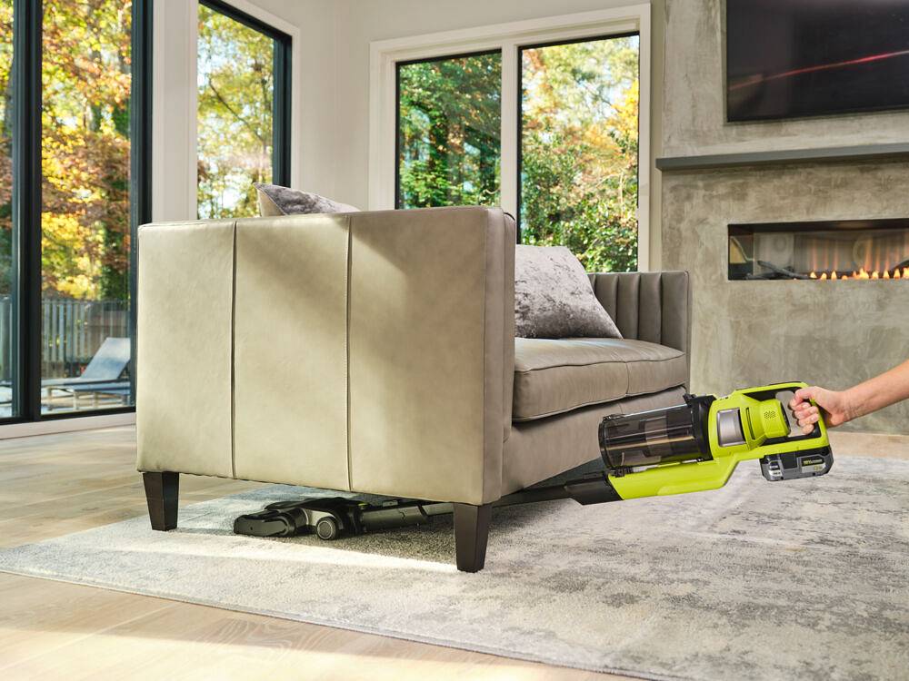 Odkurzanie dywanu pod beżową kanapą dzięki ręcznemu odkurzaczowi na akumulator RYOBI 18 V, w tle kominek i duże okna